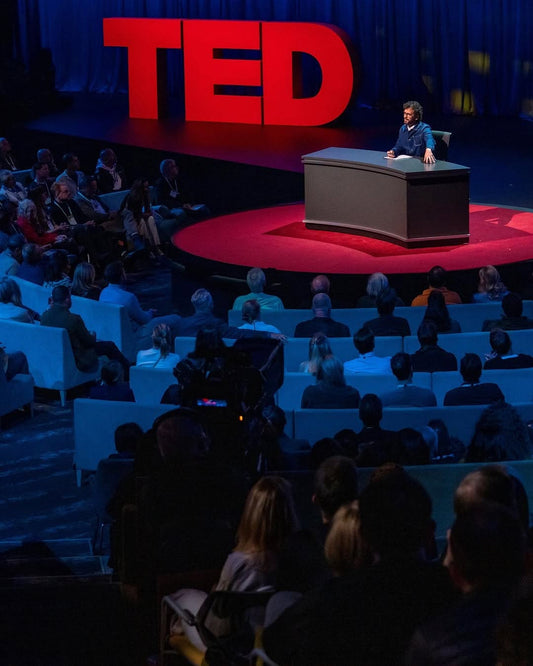 A person speaks at a TED Talk on stage with a desk, while an audience watches intently.